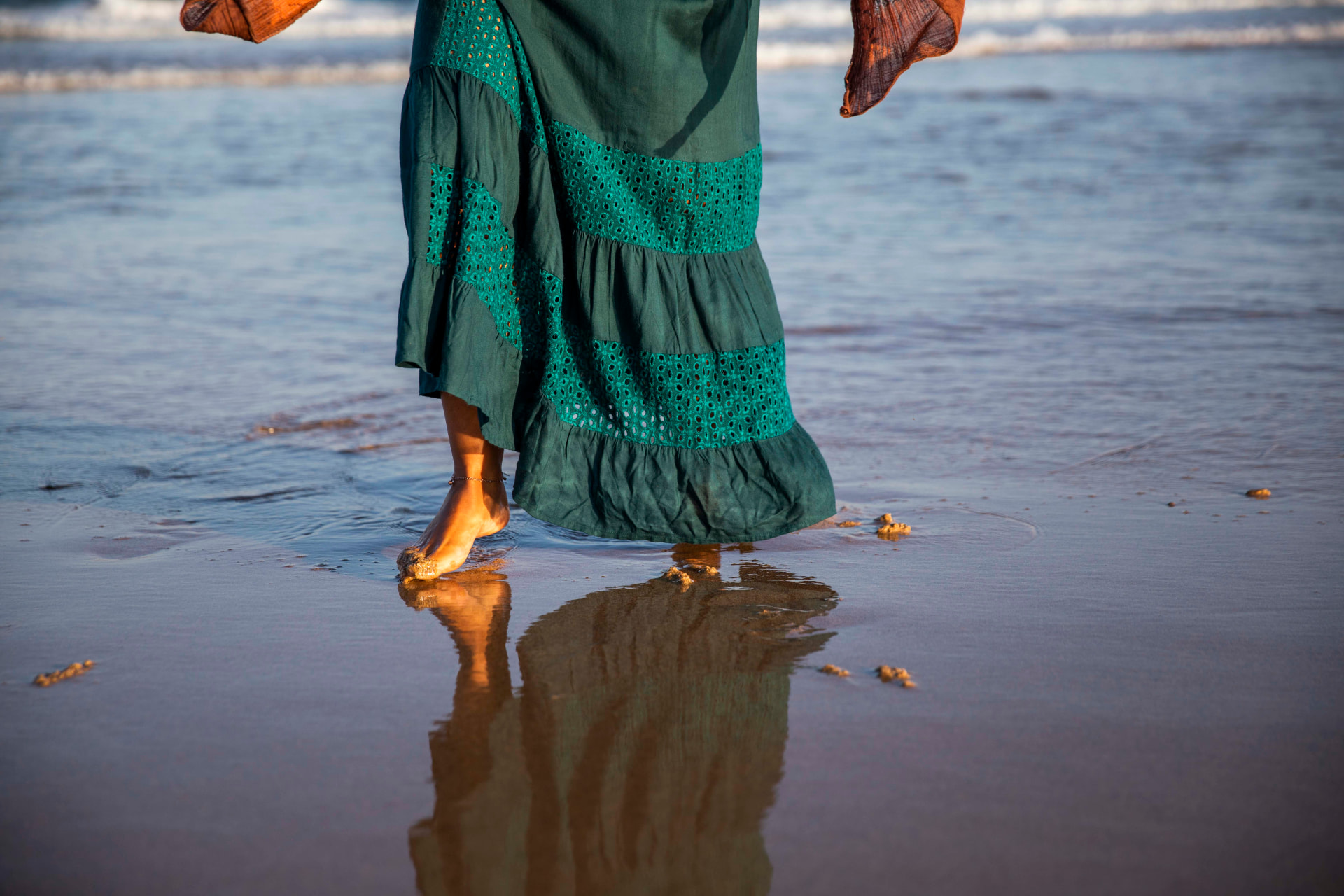 Pés descalços a caminhar na beira-mar ao pôr do sol, com vestido verde e reflexo na areia molhada